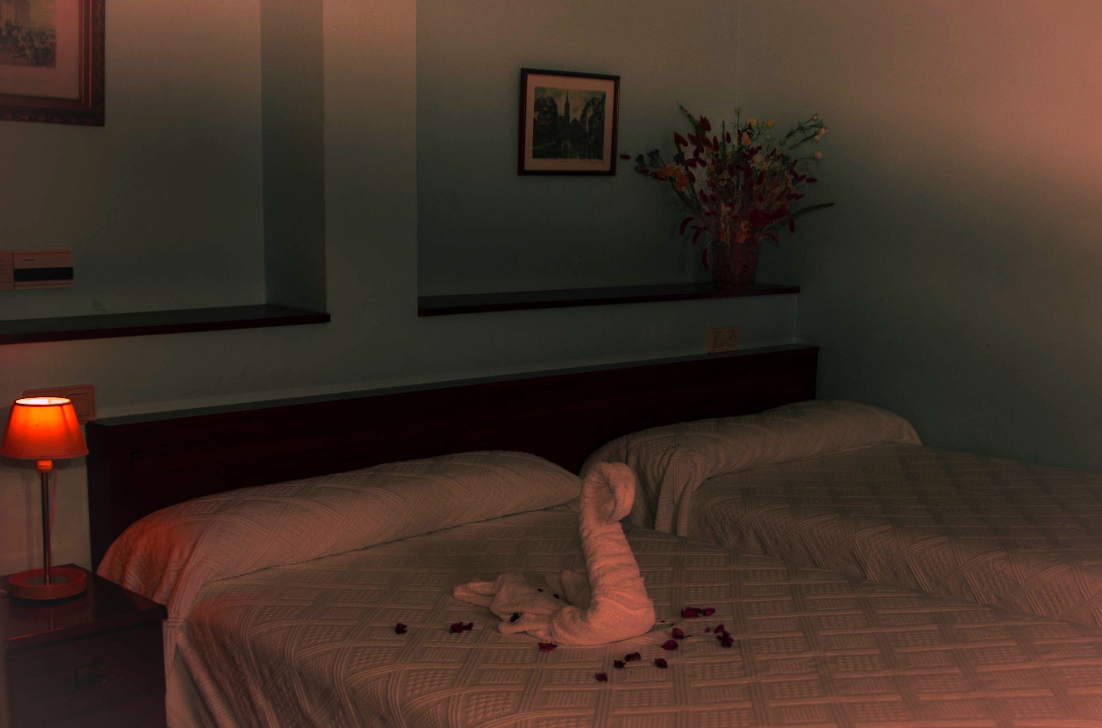 Una habitación de hotel con dos camas, una mesita de noche con lámpara, un cuadro en la pared y un florero con flores sobre una repisa.
