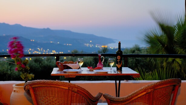 A table set for two with a bottle of wine, glasses, and a pitcher, overlooking a mountainous landscape with lights in the distance. - Elia Traditional Hotel & Spa (Platanias)