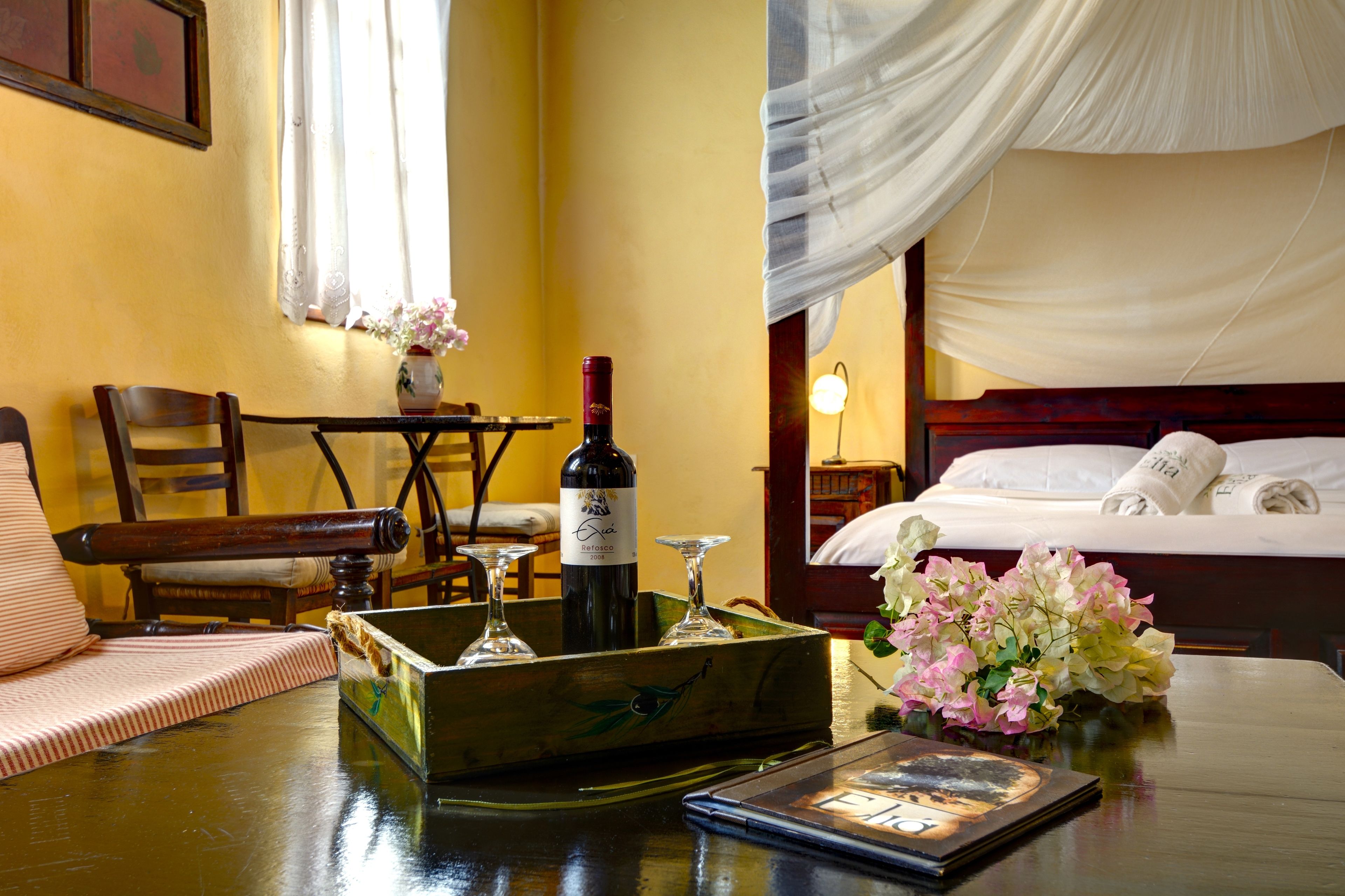 A hotel room with a bed, a table with a bottle of wine, glasses, and a vase of flowers.
