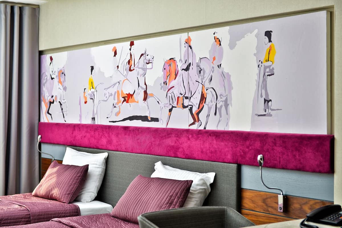 Habitación de hotel con un cuadro abstracto grande en la pared, una cama con muchos almohadones y un cabecero de madera.