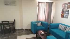 A living room with a blue sofa set, a wooden dining table with chairs, a coffee table, and a window with curtains. - Milano Hotel Suites (Amman)