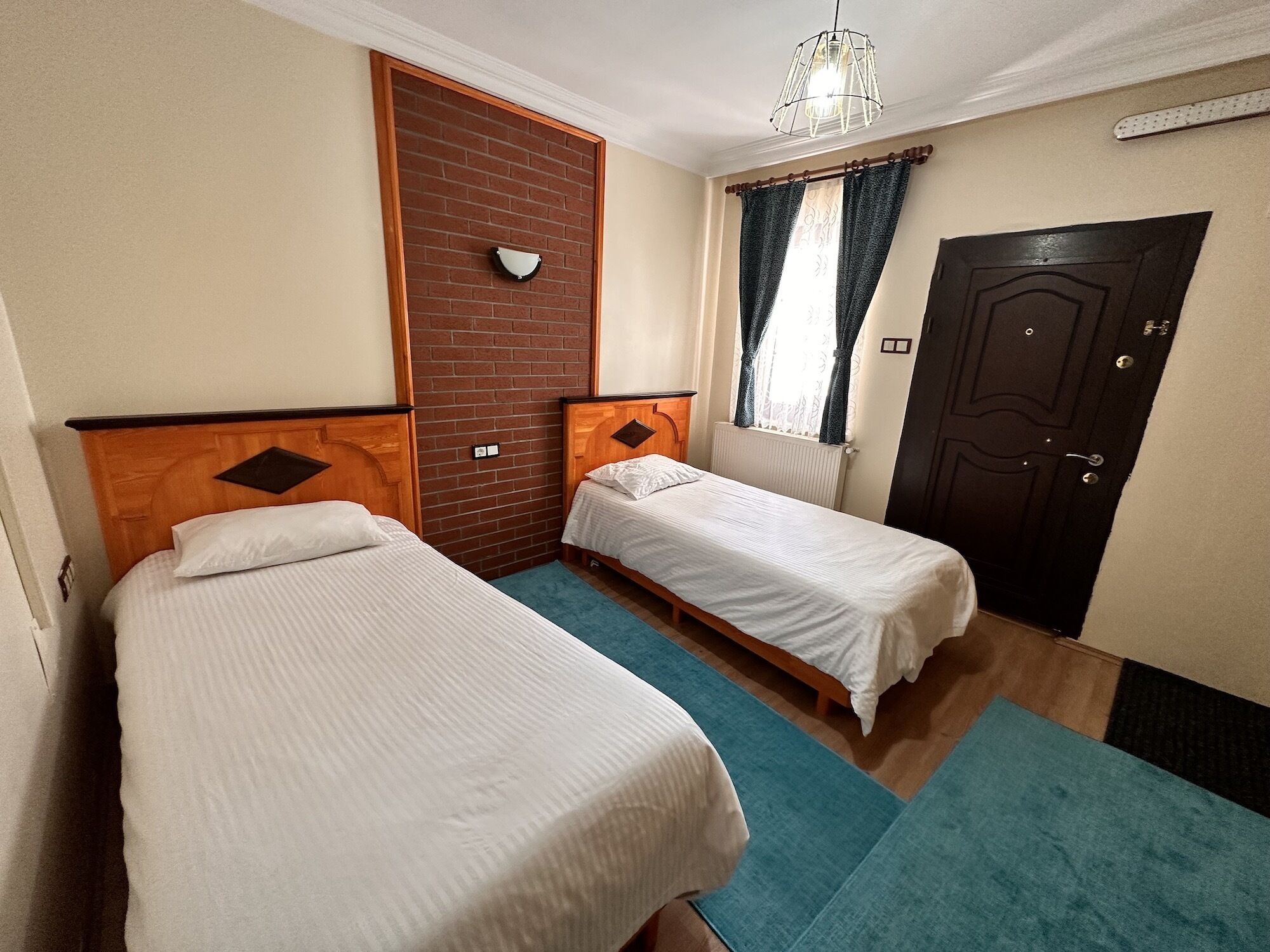 A room with two single beds, a brick partition wall, a door, and a window with curtains.