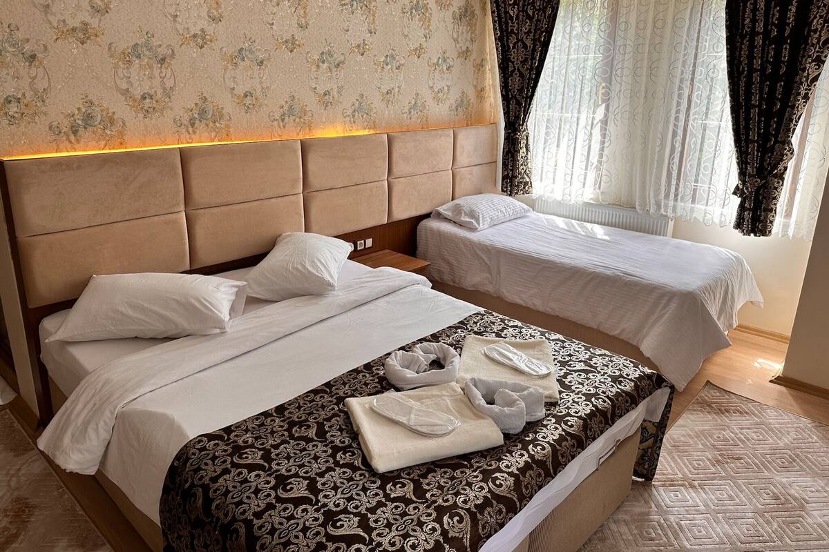 Ein Hotelzimmer mit zwei Betten, einem Kopfteil, einem Fenster mit Vorhängen und einem Badezimmerbereich mit Handtüchern.