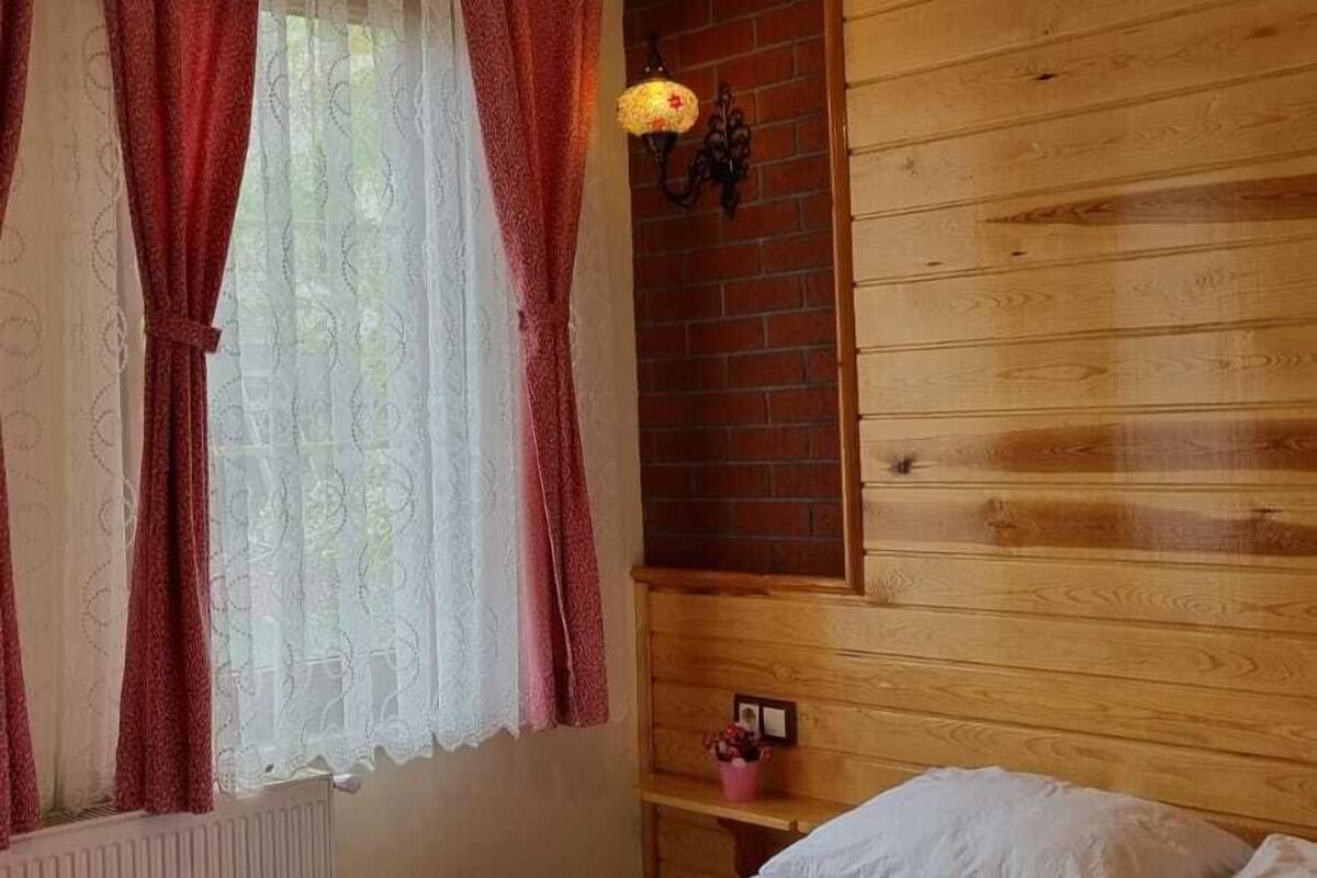 Ein Schlafzimmer mit einem hölzernen Kopfteil, einem Bett mit weißer Bettwäsche, einer Pendelleuchte und einem Fenster mit roten Vorhängen.