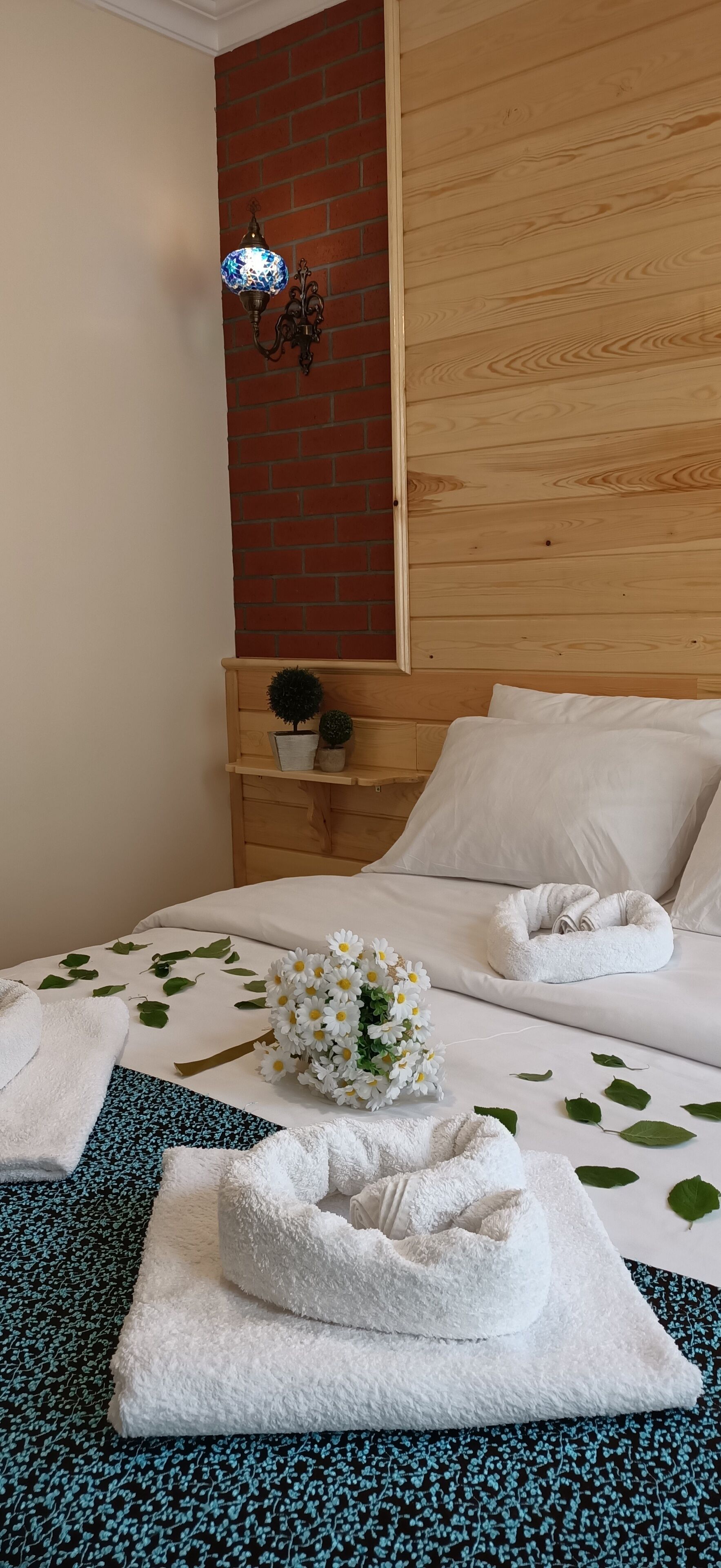 A hotel room with a wooden headboard, a brick accent wall, a nightstand with a lamp, a bed with white linens, a small potted plant, and a decorative wall sconce.