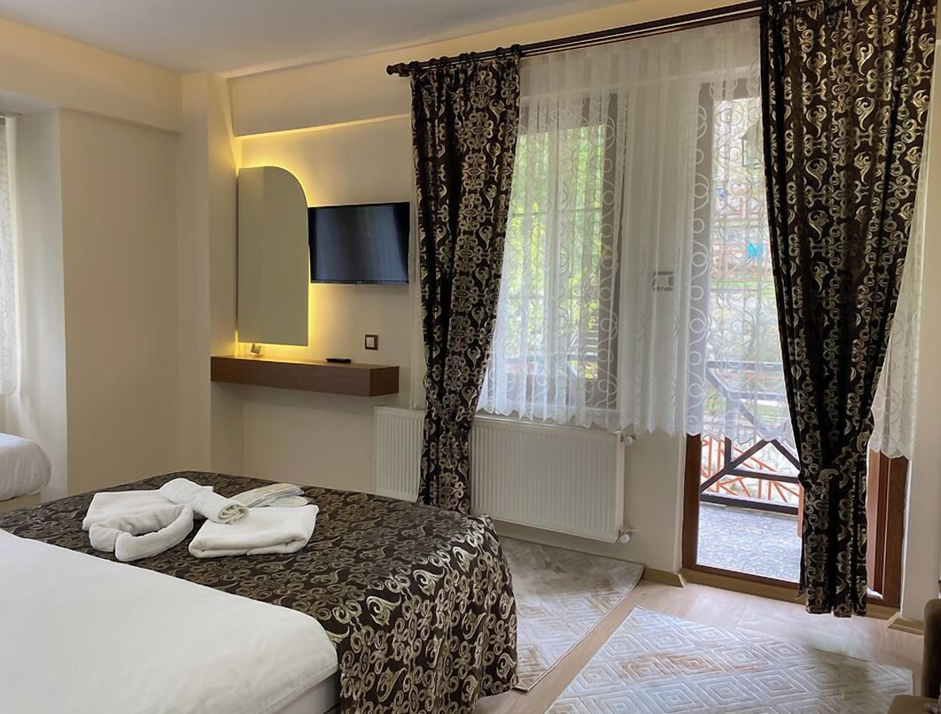 Una habitación de hotel con cama, televisor montado en la pared, ventana con cortinas estampadas y vistas al balcón exterior.