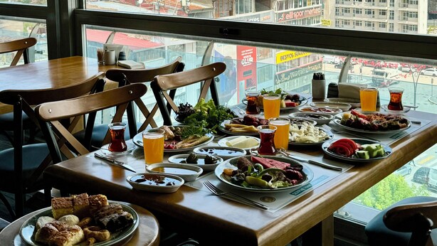 A restaurant with a table set for a meal, featuring a variety of dishes and drinks, and a view of a cityscape through large windows. - Grand Hotel Van (Van)