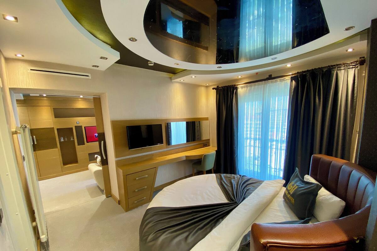 Una habitación de hotel moderna con techo curvo, cama con colcha blanco y negro, sofá de cuero, televisor montado en la pared y un amplio ventanal con cortinas.