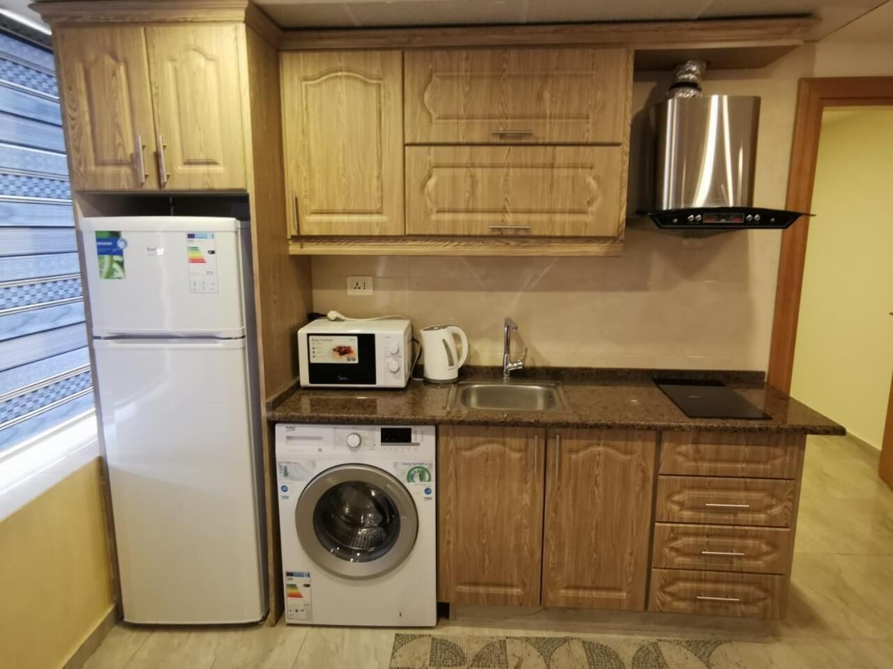 Una cocina compacta con armarios de madera, lavadora integrada, microondas y un lavamanos.