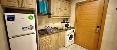 A compact kitchen with a refrigerator, microwave, washing machine, wooden cabinets, and a wooden door.