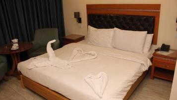 Quarto de hotel com uma cama, uma poltrona verde, uma mesinha com uma chávena e um lenço, e uma cómoda com uma lâmpada.