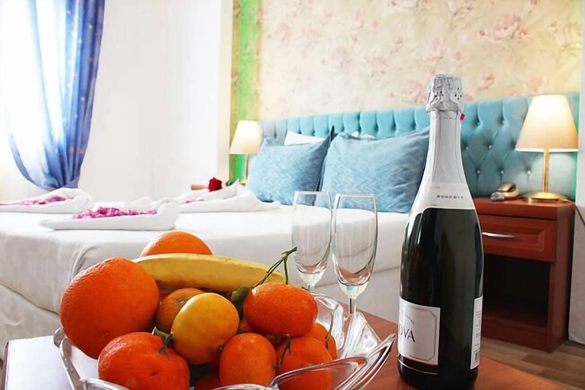 A hotel room with a bed, a bedside table with a bottle of champagne and glasses, a fruit bowl with oranges and bananas, and a blue upholstered headboard.