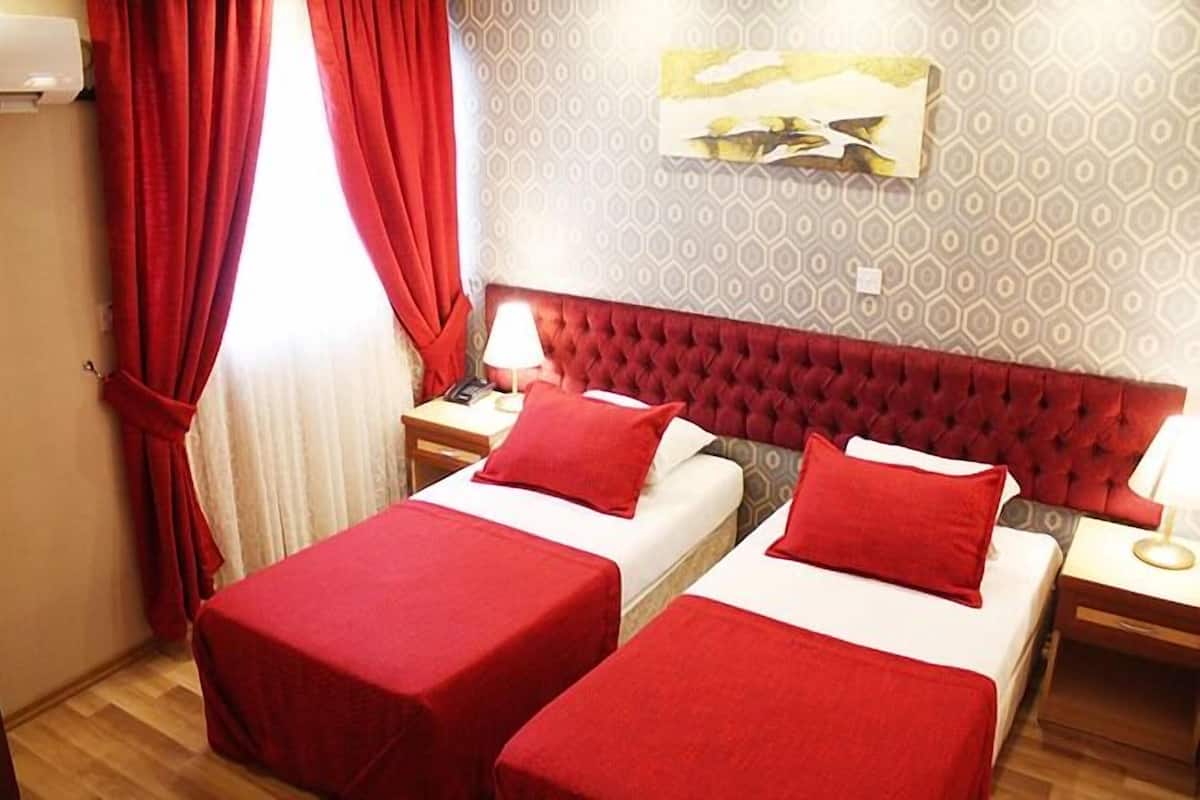 A hotel room with two beds, red bedding, a red headboard, a nightstand with a lamp, and a painting on the wall.