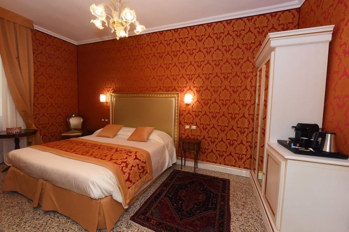 A hotel room with a large bed, a small table, a chair, a desk with a coffee maker, and a patterned carpet.