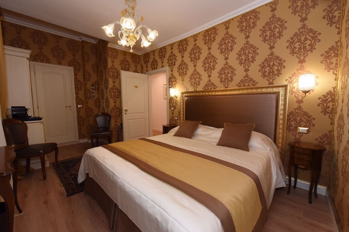 A hotel room with a large bed, a desk, and a chair. There is a chandelier above the bed and a lamp on the bedside table.