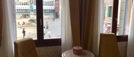 Una habitación con dos sillas y una mesa pequeña, una ventana que da a un edificio y una estatua, y una cortina entreabierta.