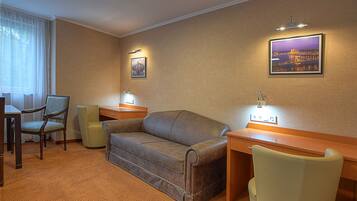 A hotel room with a sofa, two armchairs, a desk, and a chair. There are two wall-mounted lamps and two framed pictures on the wall.