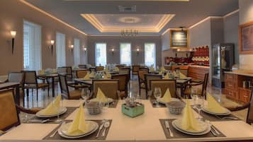 A spacious dining area with multiple tables set for service, a kitchen in the background, and a modern ceiling design.
