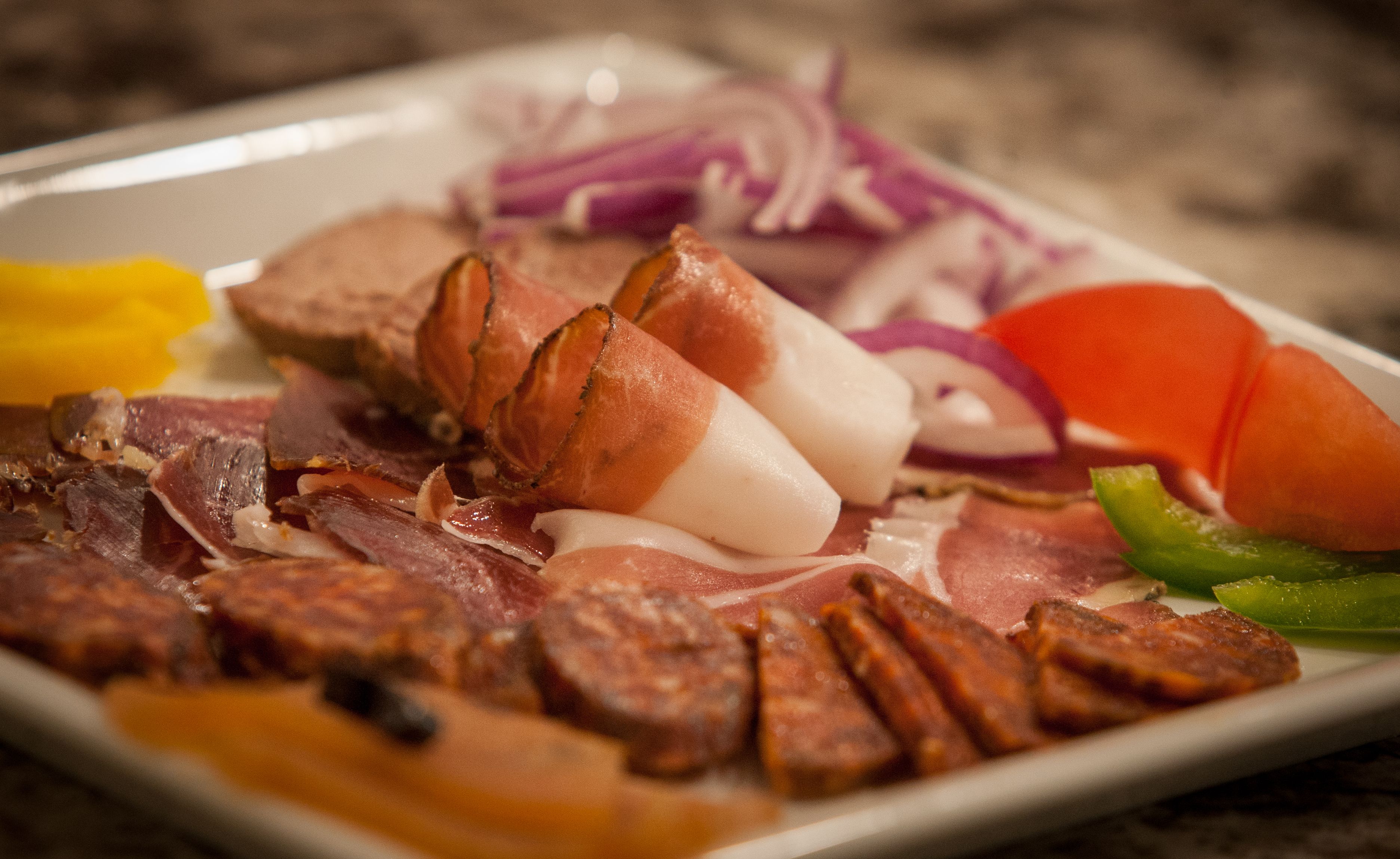 A plate of assorted meats including bacon, ham, and possibly prosciutto, accompanied by sliced vegetables like red onions and carrots.