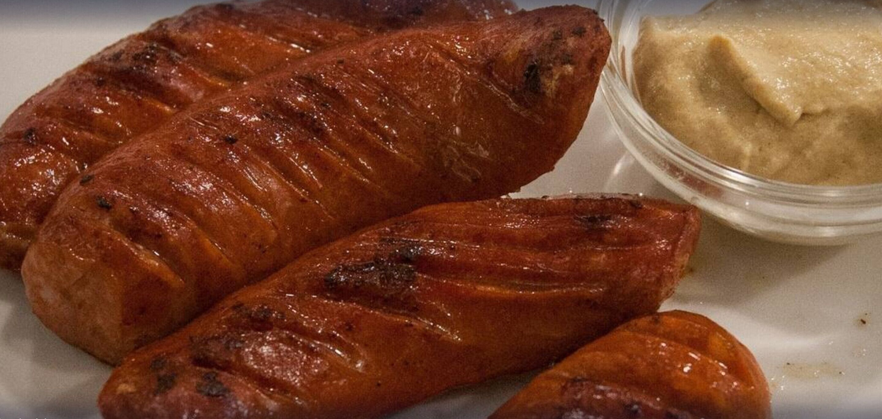 Three grilled sausages and a small container of mustard on a white plate.