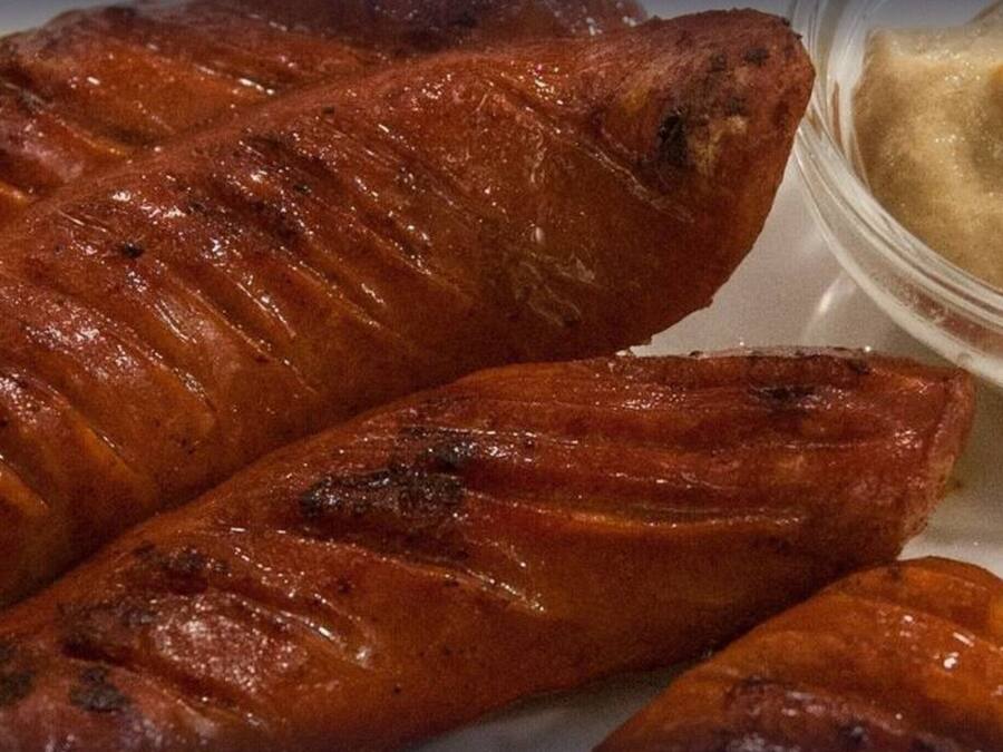 Tres salchichas a la parrilla y un recipiente pequeño de mostaza sobre un plato blanco.