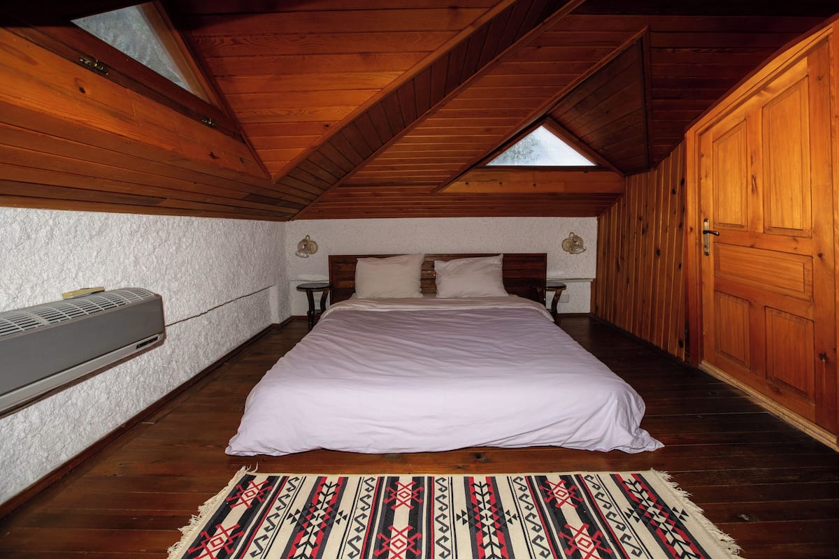 A bedroom with a wooden ceiling, a bed with white bedding, a patterned rug, and a wall-mounted air conditioner.