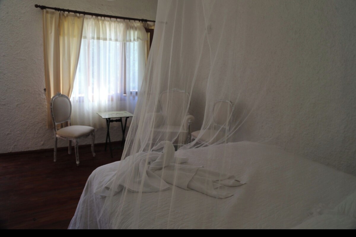 Ein Schlafzimmer mit einem Bett, einem Sessel, einem kleinen Tisch und einem Fenster mit Vorhängen.