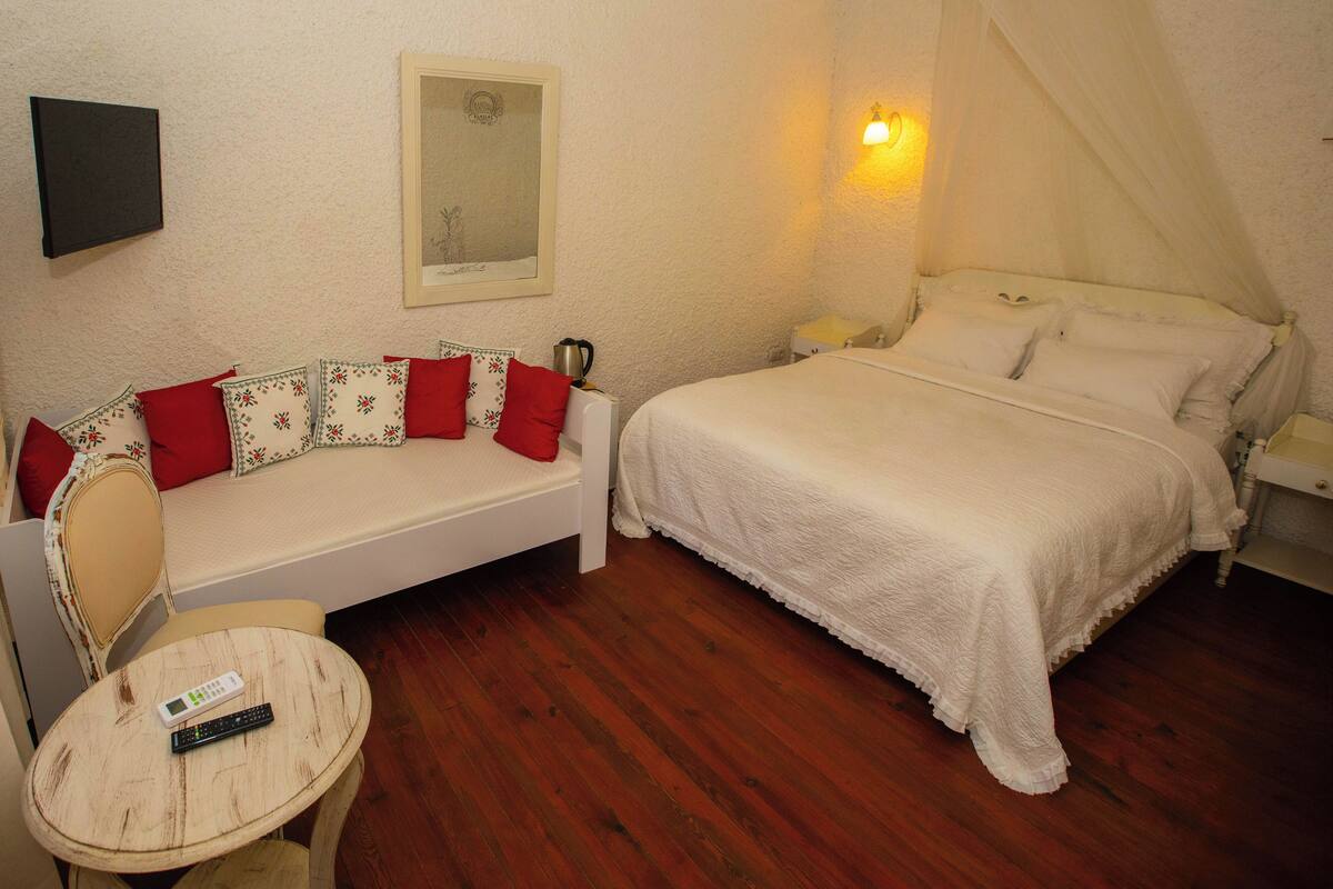 A bedroom with a bed, a sofa, a small table, a remote control, and a wall-mounted light fixture.