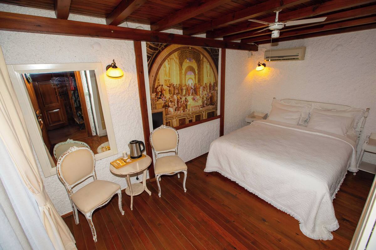 A bedroom with a large bed, a chair, a small table with a kettle, and a painting on the wall.