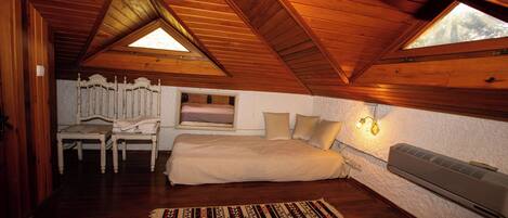 Un dormitorio con techo inclinado, una cama, una silla, un ventanal y un calentador fijado a la pared.