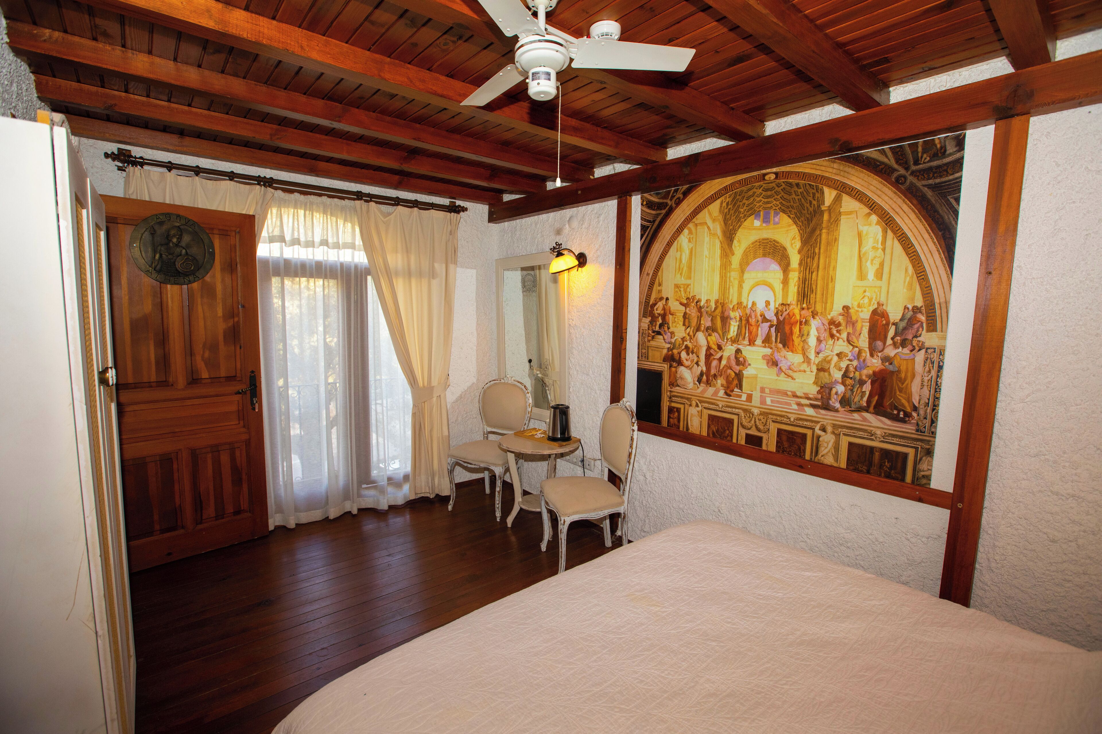 A room with a bed, a ceiling fan, a dining table with chairs, a painting on the wall, and a wooden door.
