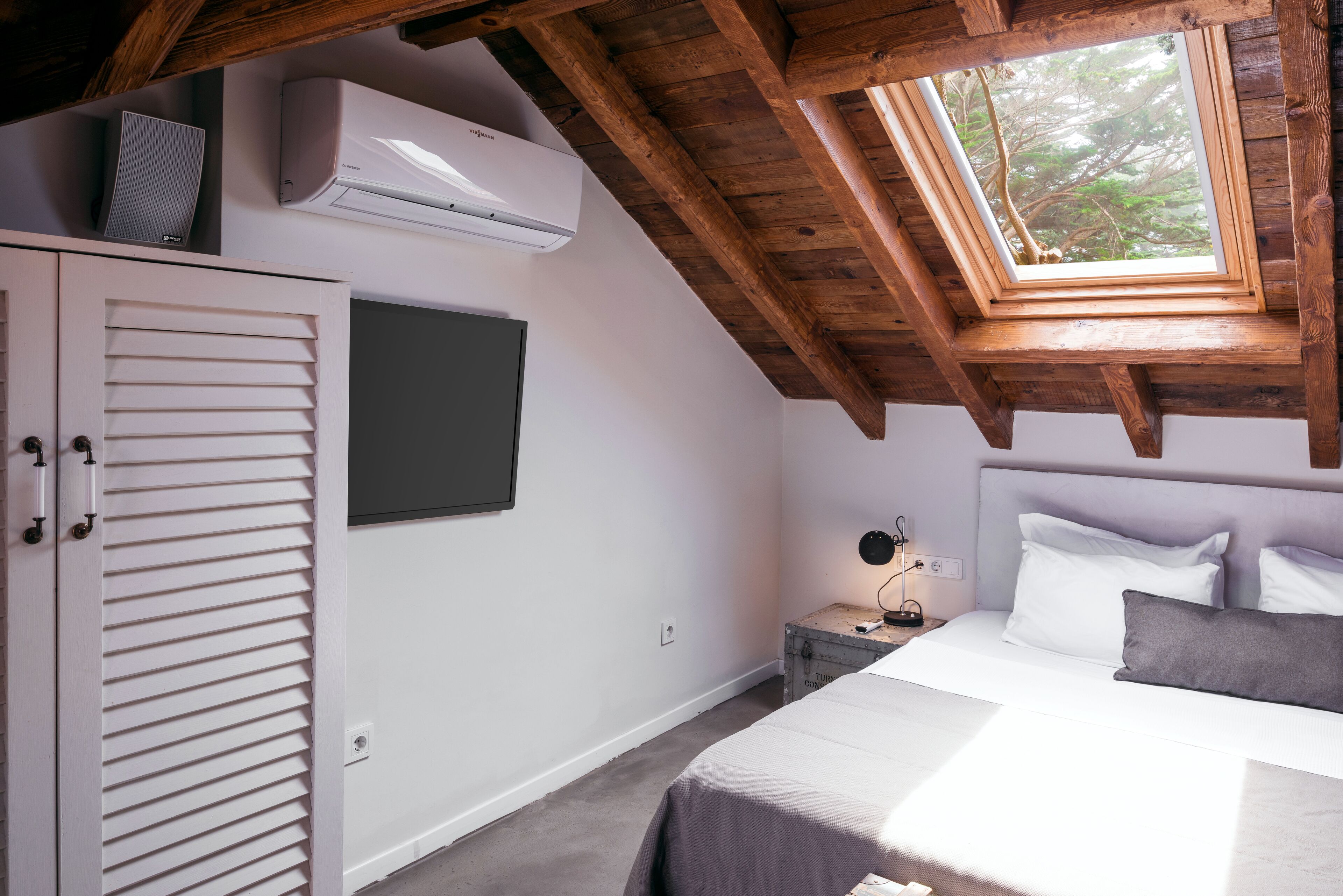Un dormitorio con techo inclinado, un tragaluz, una cama con almohadas, una mesita de noche con lámpara y un armario con persianas horizontales.
