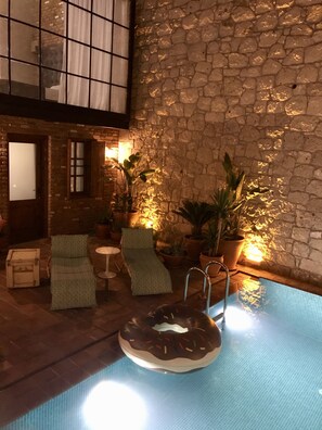 An indoor swimming pool with a donut-shaped float, lounge chairs, and potted plants against a stone wall.