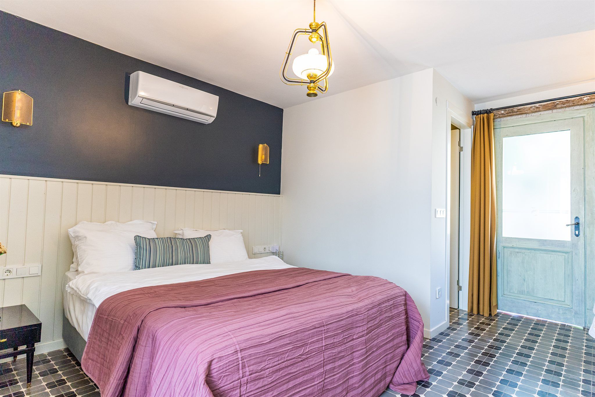 Hotellihuone, jossa on sänky, yöpöydät, ilmastointilaite, seinään kiinnitetty valaisin ja ovi, josta on näkymä ulos.