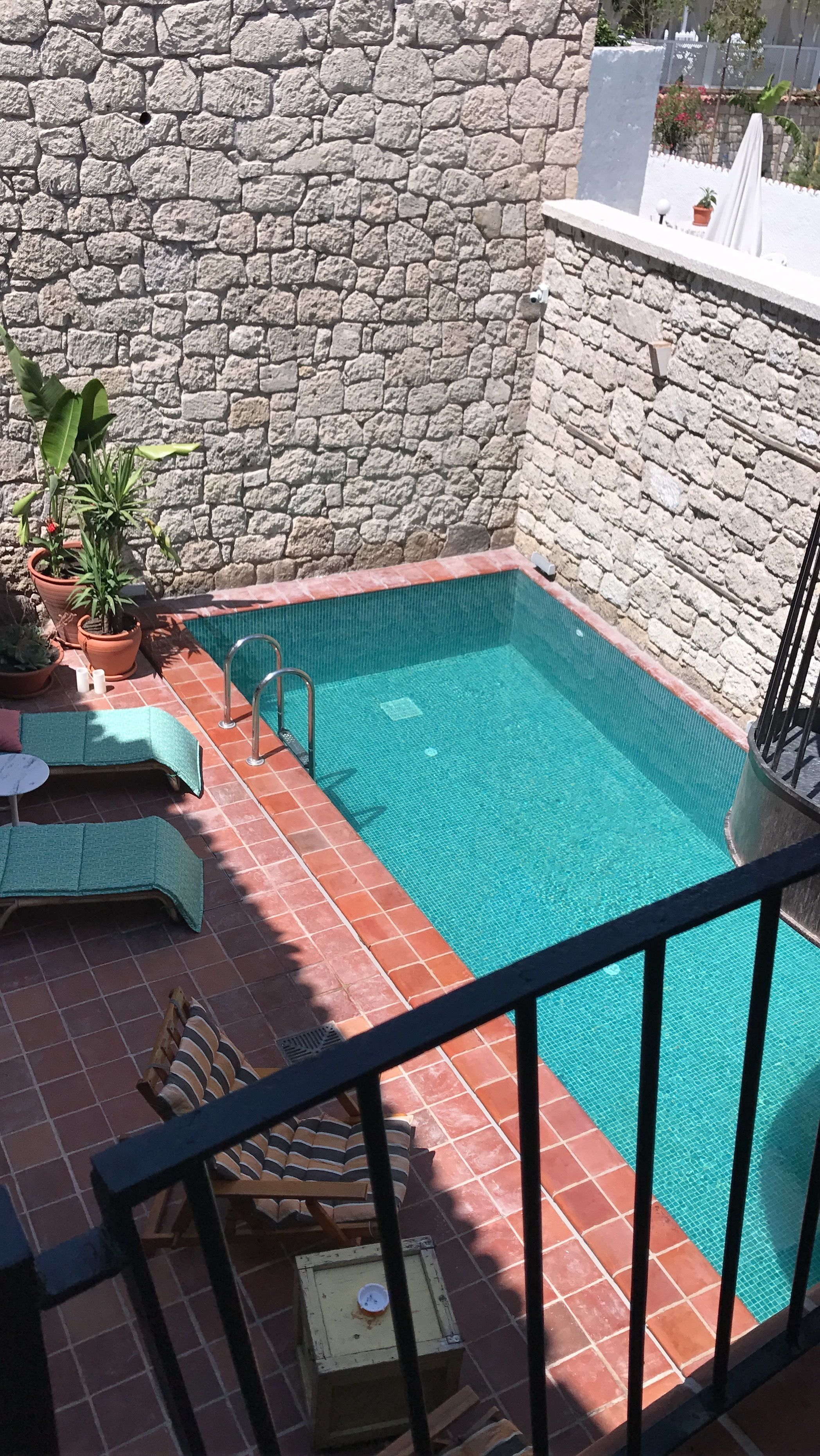 A small outdoor swimming pool with a stone wall backdrop, a tiled deck, and a few potted plants.