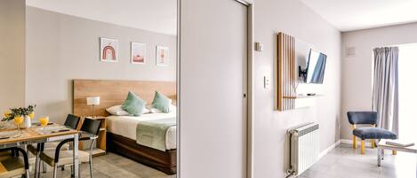 A modern hotel room with a bed, a dining table, chairs, a TV, and artwork on the wall.