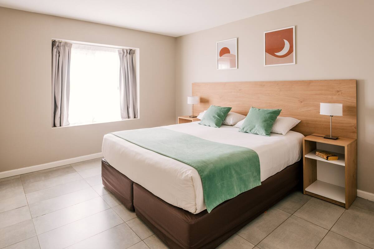 A bedroom with a large bed, two bedside tables, a window with curtains, and two wall-mounted pictures.