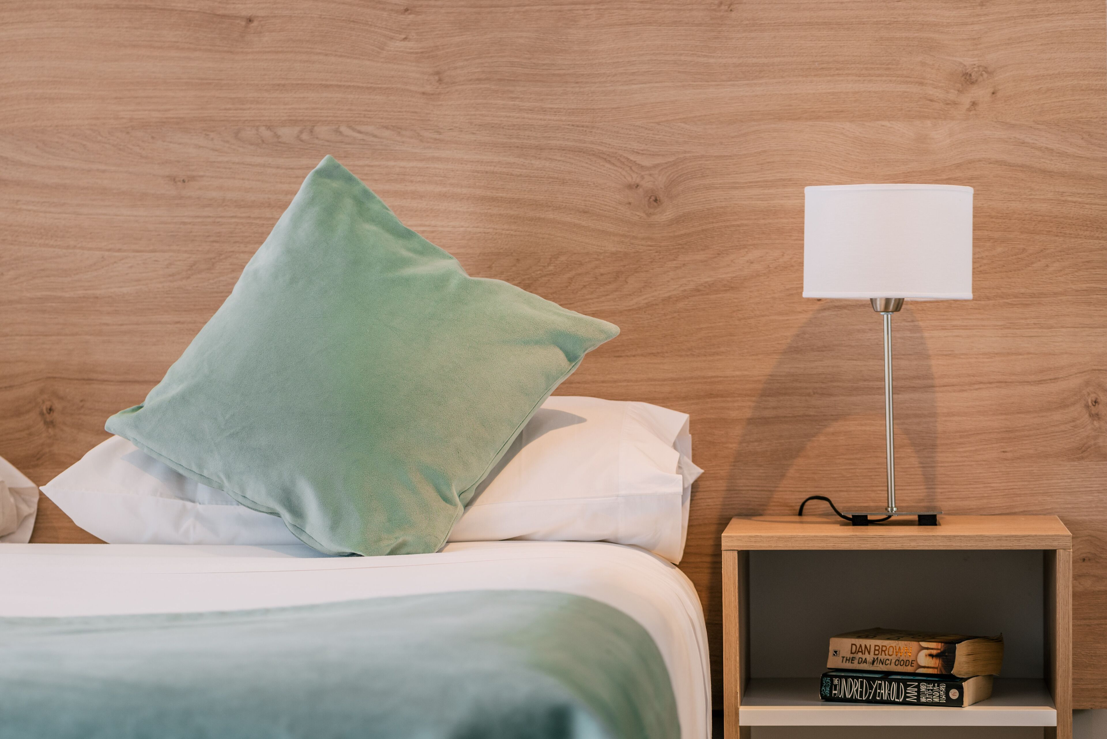 Cama bem arrumada com um travesseiro verde, um travesseiro branco e um edredom branco. Um criado-mudo com uma luminária e alguns livros. A parede atrás da cama é de madeira.