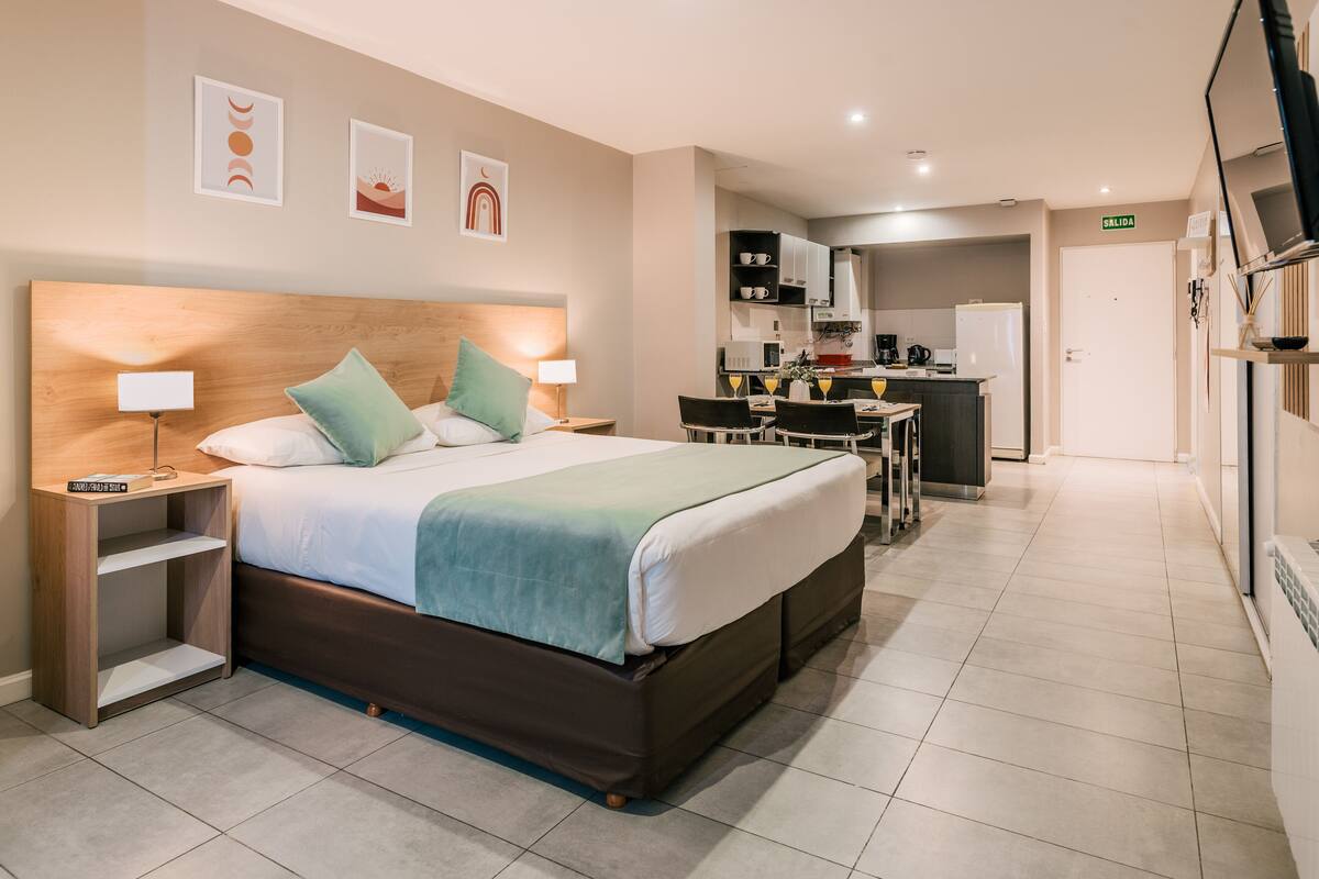 A hotel room with a large bed, bedside tables, a desk, and a kitchenette in the background.