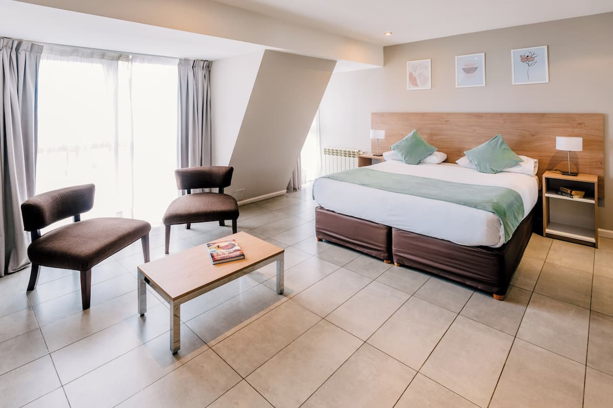 Una habitación de hotel con cama, dos sillones, una mesita, una mesita de noche y un ventanal con cortinas.