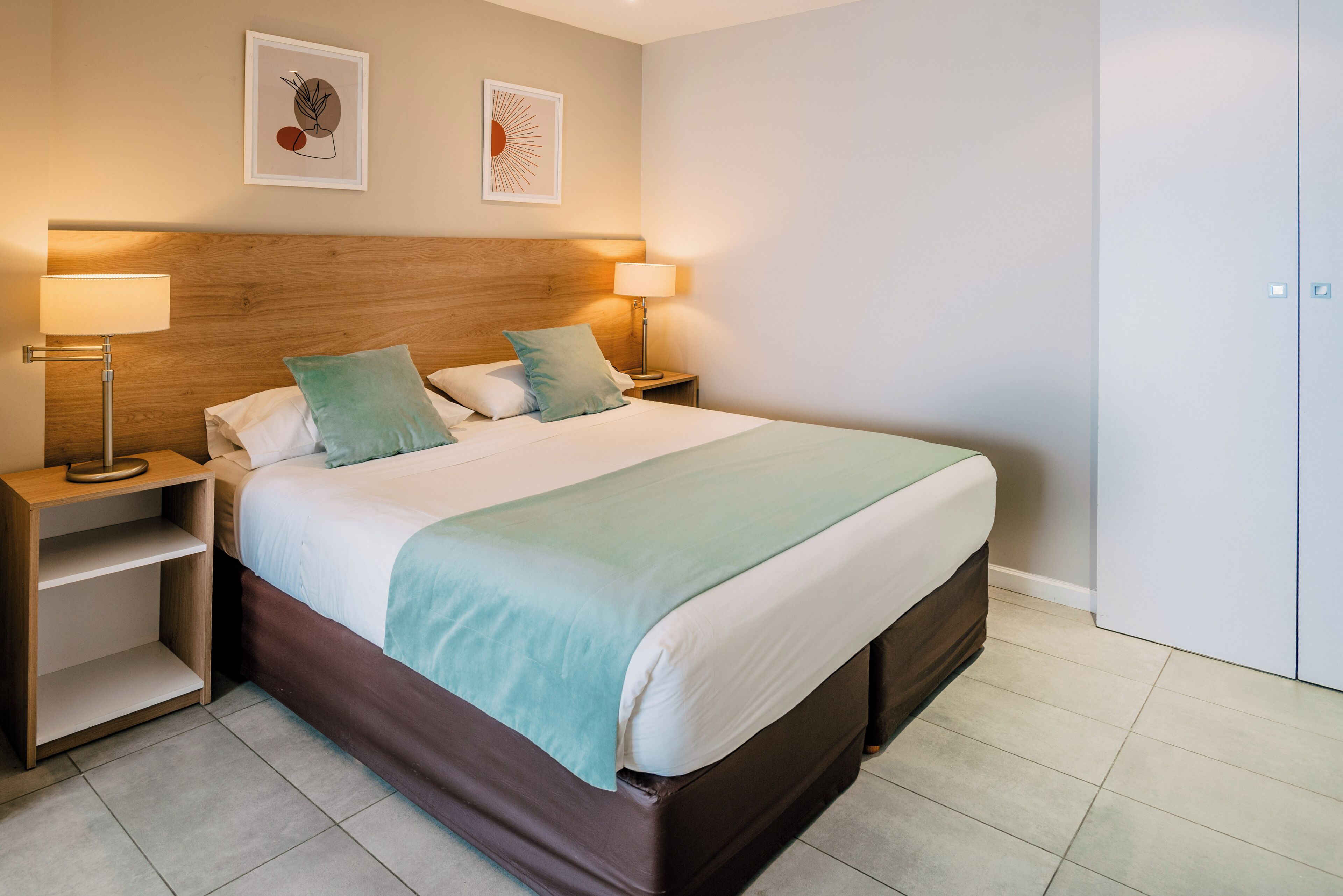 A neatly made bed with a wooden headboard, two bedside lamps, and a nightstand with a shelf.