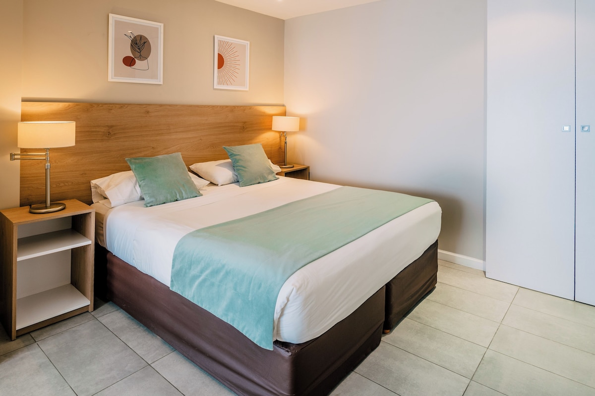A neatly made bed with a wooden headboard, two bedside lamps, and a nightstand with a shelf.