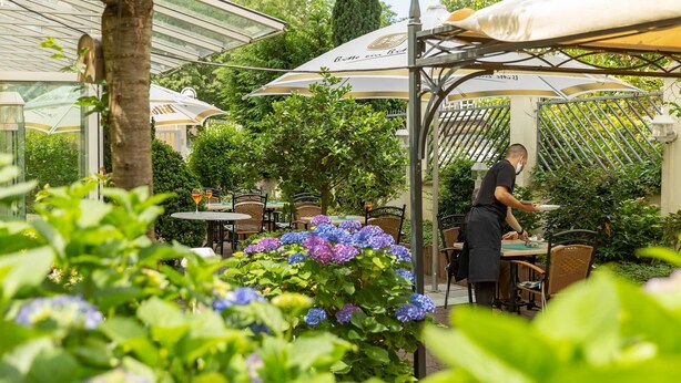 Un café en plein air avec des tables et des chaises sous des parasols, entouré d’une végétation luxuriante et de fleurs.