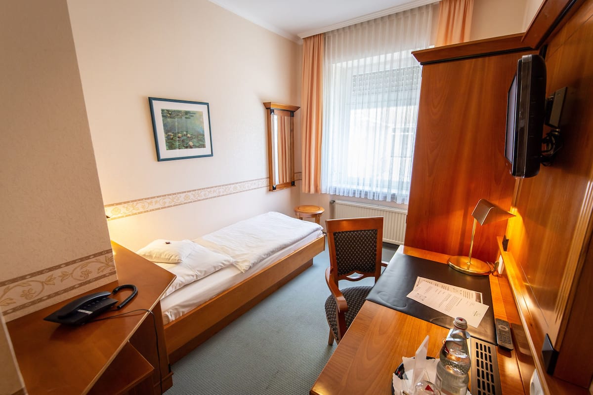 Ein Hotelzimmer mit einem Einzelbett, einem Schreibtisch mit Stuhl, einem Telefon, einem Fernseher und einem Fenster mit Vorhängen.