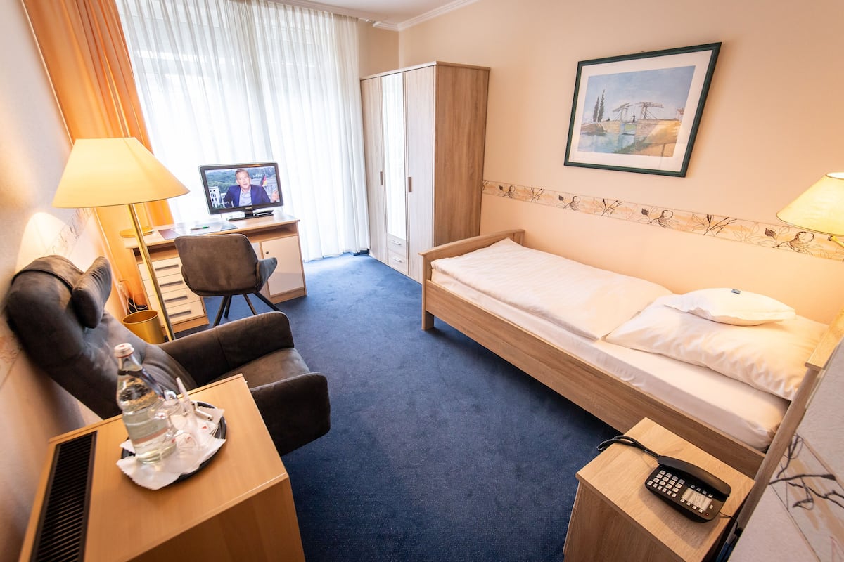 Ein Hotelzimmer mit einem Einzelbett, einem Schreibtisch mit Computer, einem Stuhl, einem Kleiderschrank und einem Nachttischtelefon.