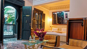 Habitación de hotel con cama, zona de estar con una mesa de vidrio, una silla y un jarrón decorativo con flores.