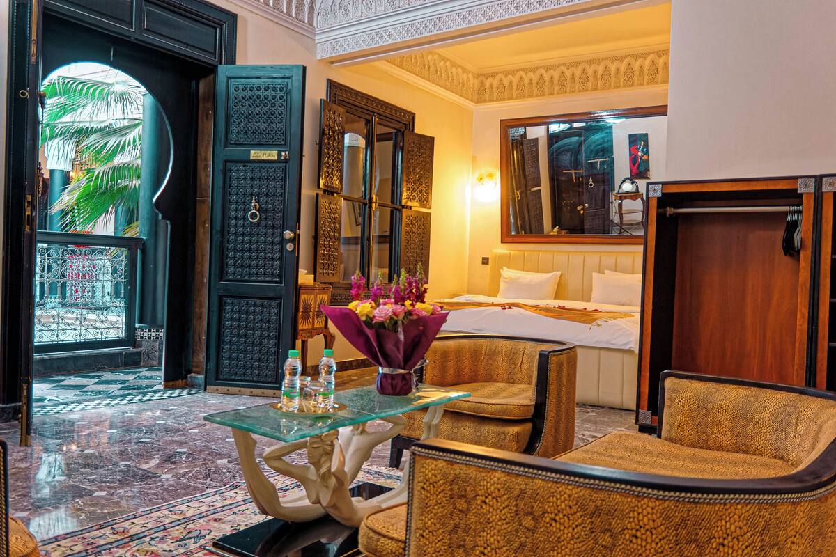 Habitación de hotel con cama, zona de estar con una mesa de vidrio, una silla y un jarrón decorativo con flores.