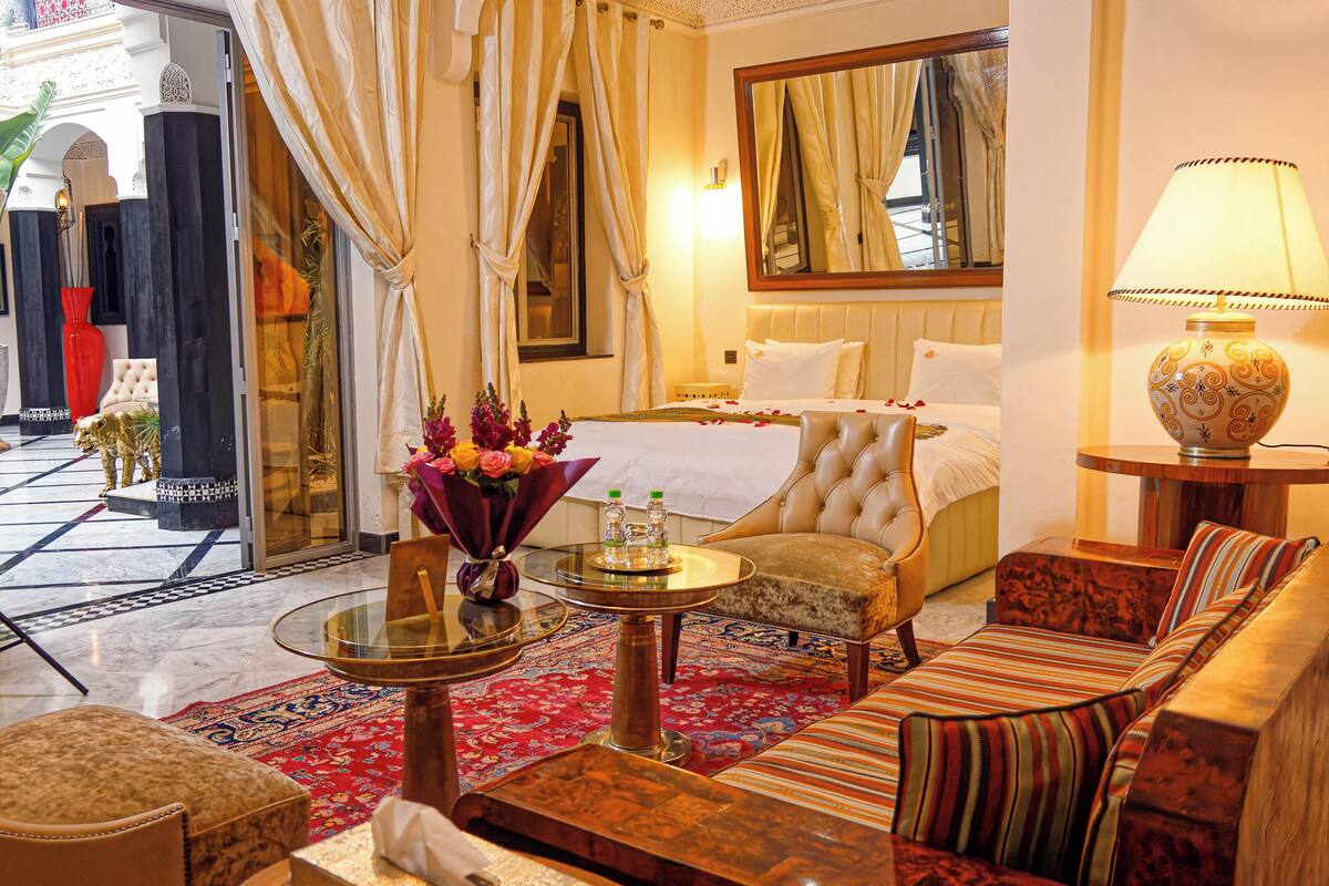 Habitación de hotel con una cama, zona de estar con un sofá y sillones, una mesita con un jarrón de flores y una lámpara.