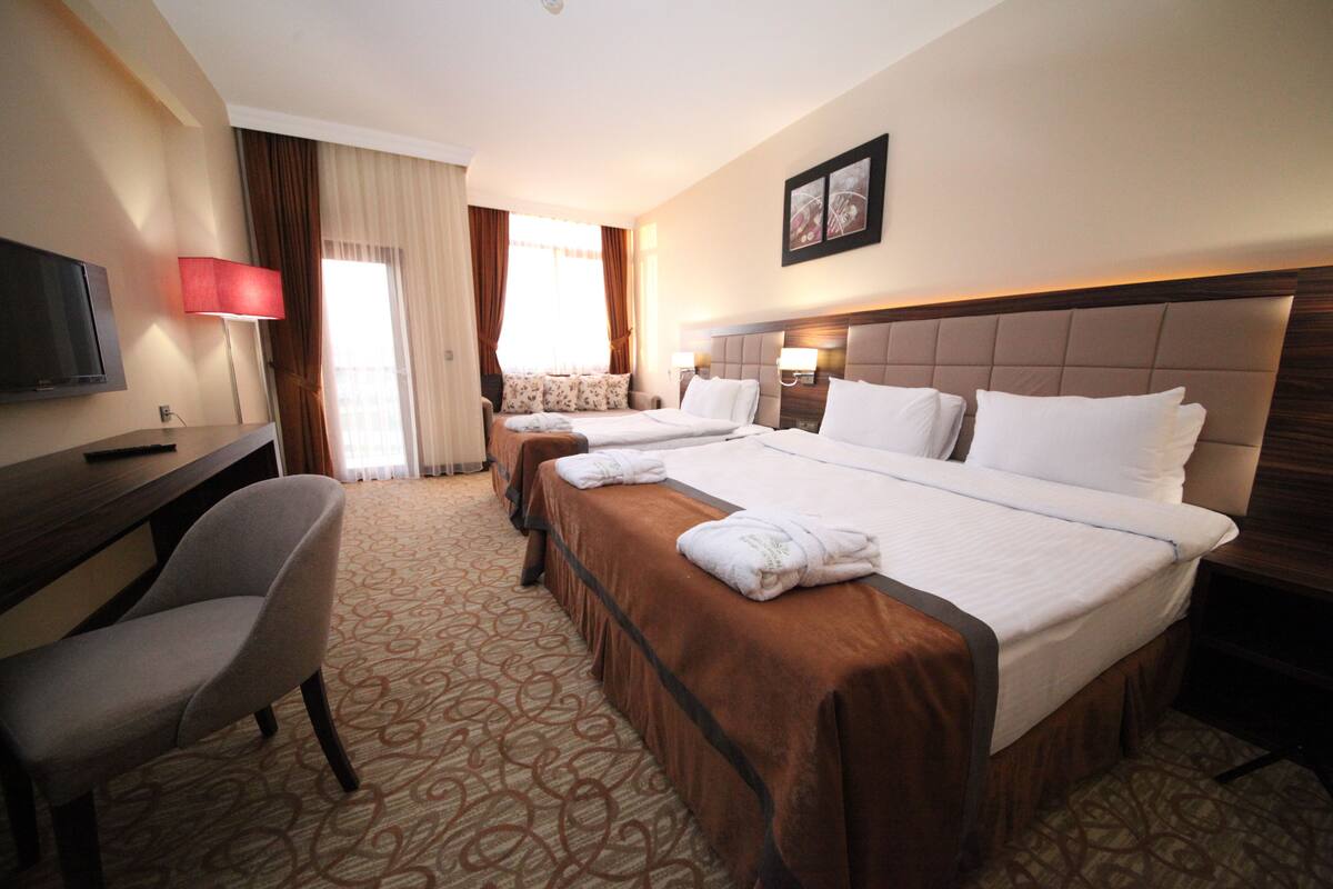 A hotel room with a large bed, a smaller bed, a desk with a chair, a TV, and a window with curtains.