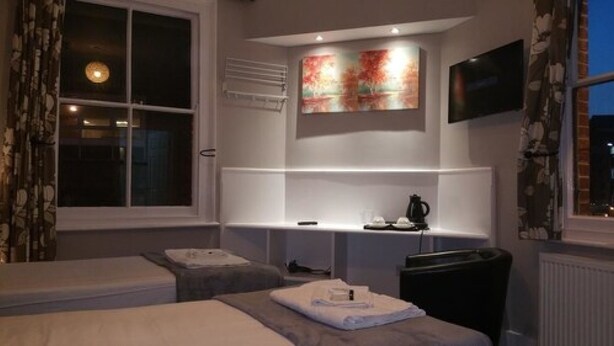A hotel room with a bed, a bedside table, a TV mounted on the wall, and a window with curtains. - Ali Inn (Oxford)